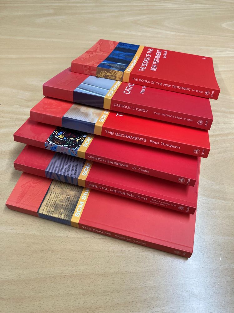 A stack of 7 red SCM Studyguide books on a table, including The Sacraments, Catholic Liturgy, Biblical Hermeneutics and The Books of the New Testament