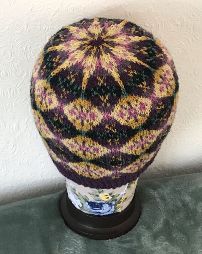 A hand knitted hat in stranded colourwork, showing a diamond pattern, mainly in shades of purple and yellow. 
The top has a 9 pointed star where the  stitch count has been reduced to form the crown. 
