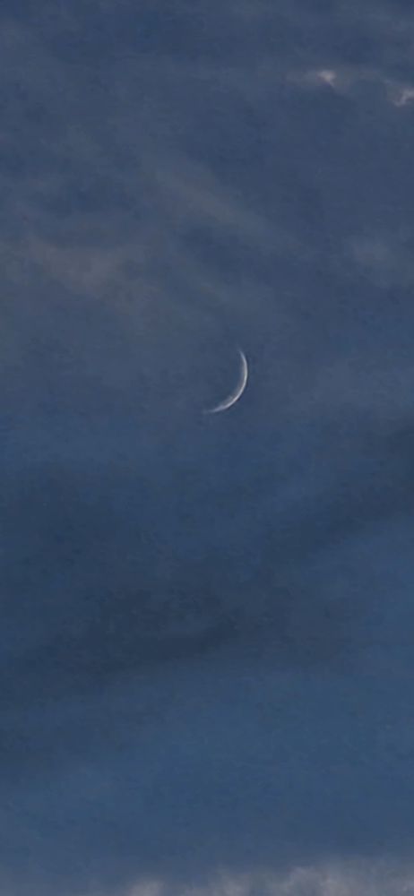 A very slim waxing crescent moon behind thin altostratus clouds
