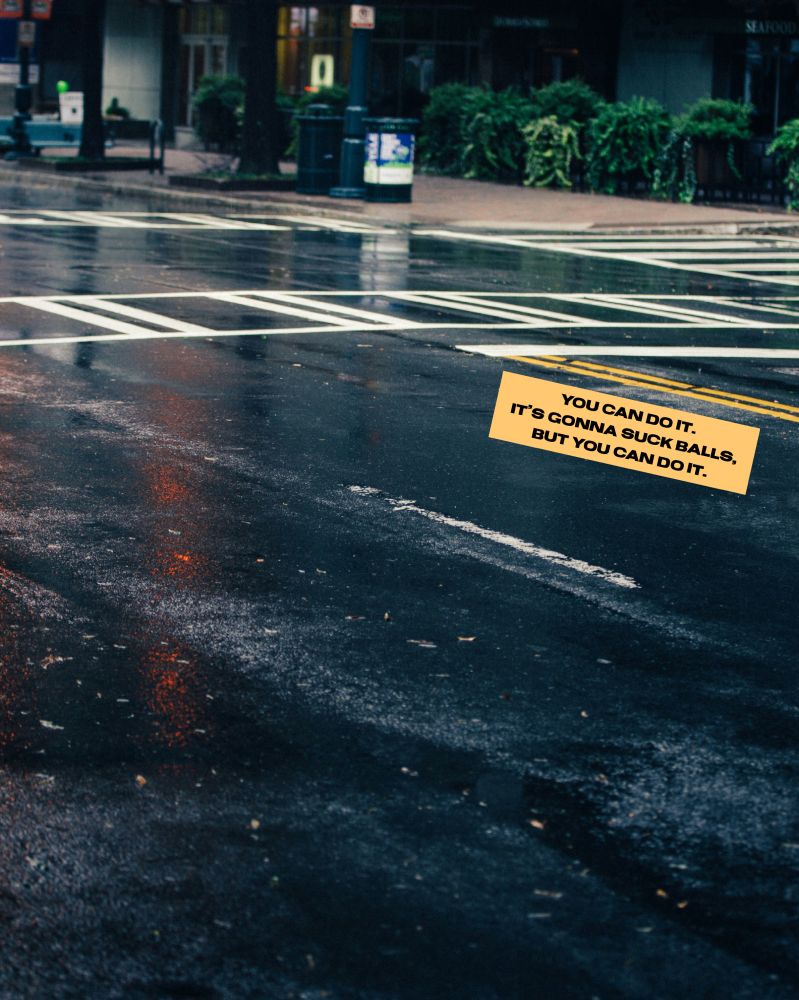 On a photo of a rainy city street, a small yellow text box is aligned on an angle with the double yellow stripes on the road. It reads, “You can do it. It’s gonna suck balls, but you can do it.”