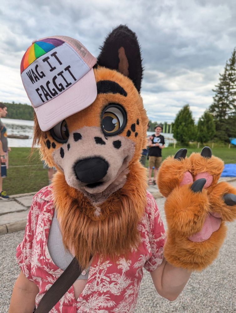 Canine fursuit wearing a red shirt and a "Wag It Faggit" shirt, making the peace sign with their left paw. Taken at Camp Feral, Ontario, Canada.