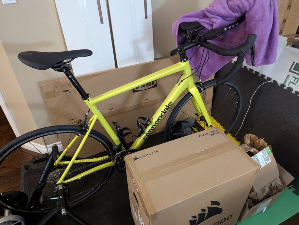 A yellow bike set up, surrounded by various boxes (an IKEA box in the background and a computer case in the foreground).