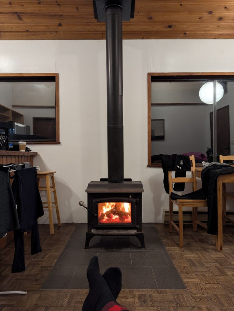 Fire burning in a wood stove in a small cabin. Wet clothes are draped on a drying rack and nearby chairs.