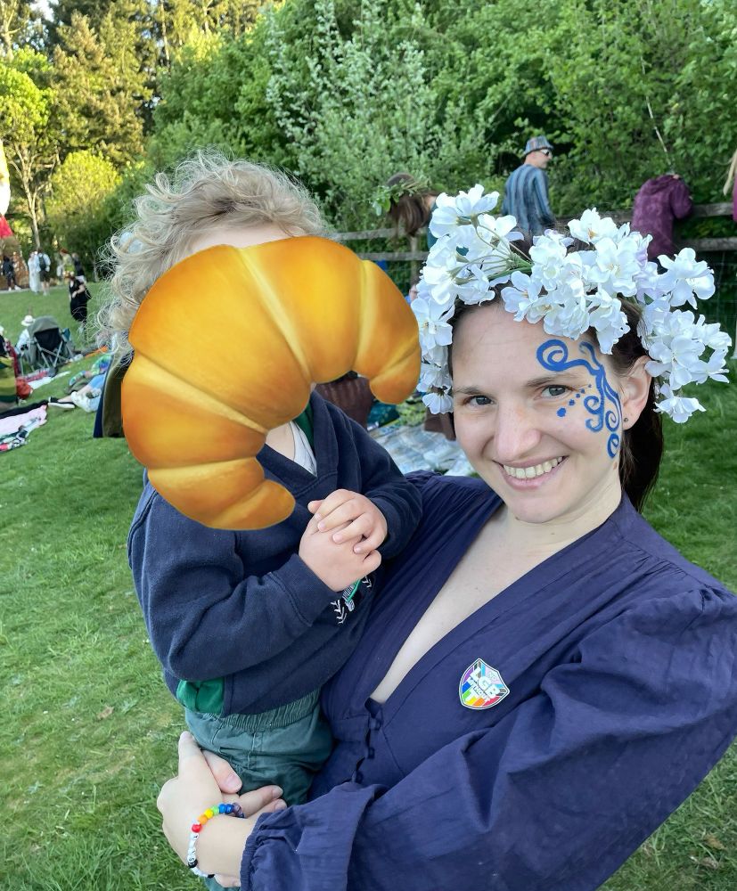 Me, posing wearing a blue dress and face paint, holding a toddler whose face is hidden with a croissant emoji 