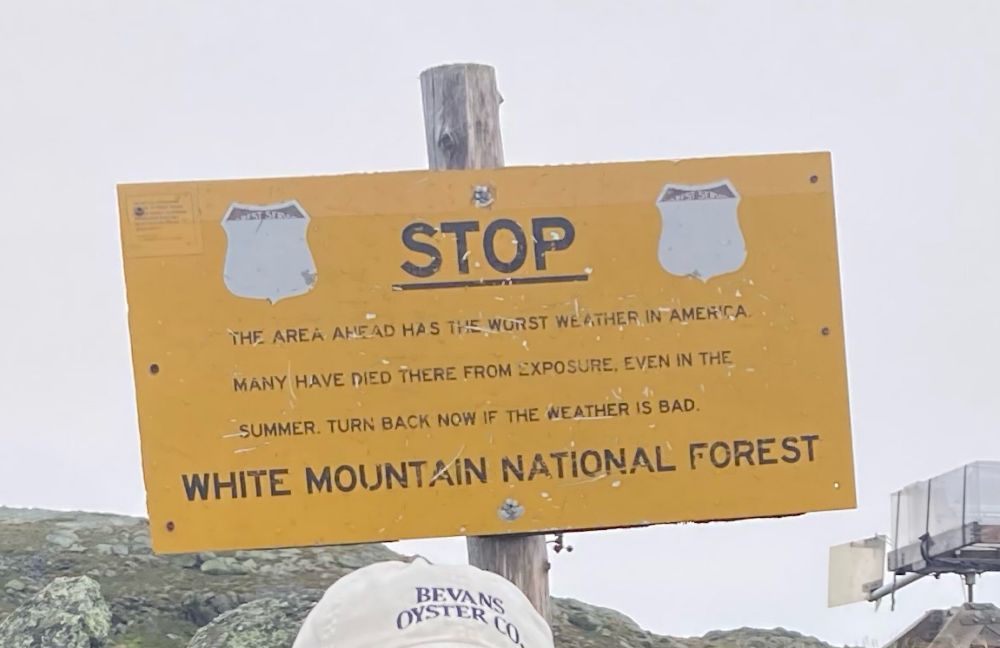 Battered yellow Forest Service sign that says “STOP. The area ahead has the worst weather in America. Many have died there from exposure even in the summer. Turn back now if the weather is bad. White Mountain National Forest.”