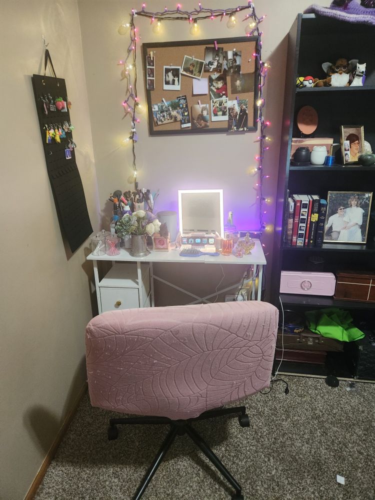 A vanity setup featuring a white desk with drawers, a pink upholstered chair, and various makeup items and decorations on the desktop. A small mirror is illuminated by soft purple and white LED lights. Above the desk, a corkboard is covered with polaroid photos and surrounded by multicolored string lights. To the right is a dark bookshelf holding books, framed photos, two cat urns, one black(Bart) and one white(Hekate) and decorative items. The area has a warm and personalized aesthetic
