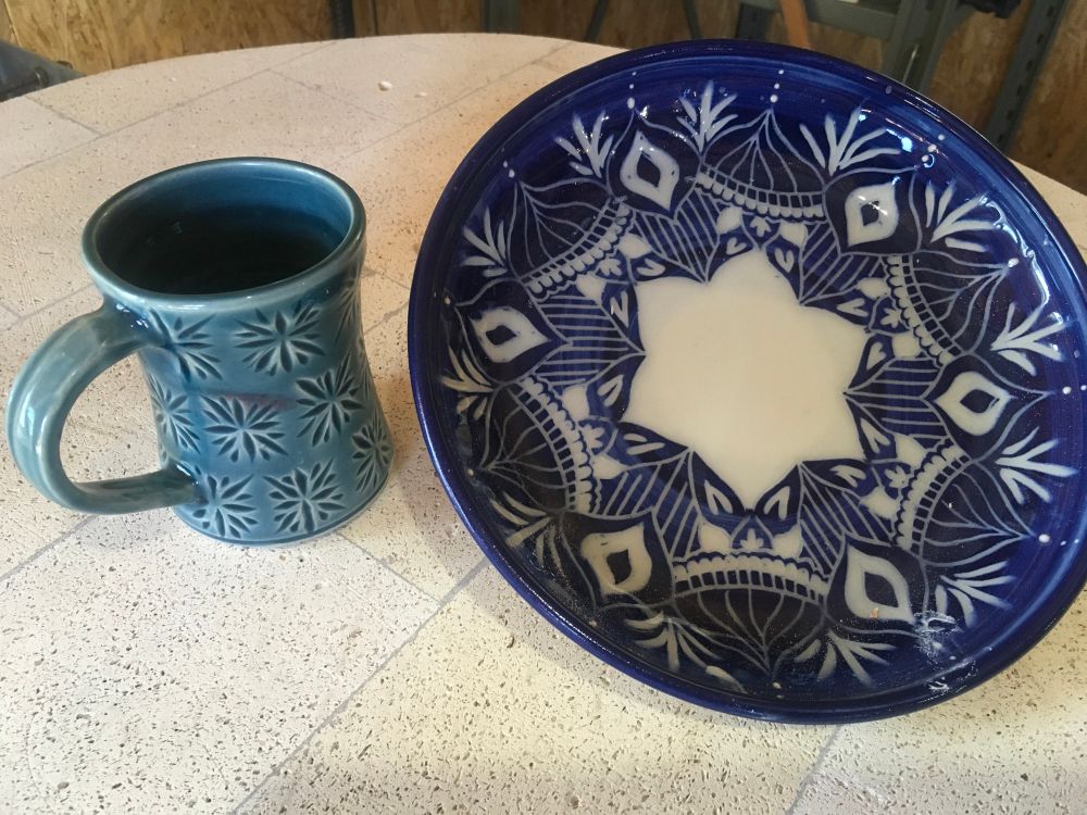 Photo of a handmade carved mug in teal, and a shallow rimless plate with a central 6 pointed white area, with the remainder painted in cobalt blue slip and carved in a mandala pattern.
