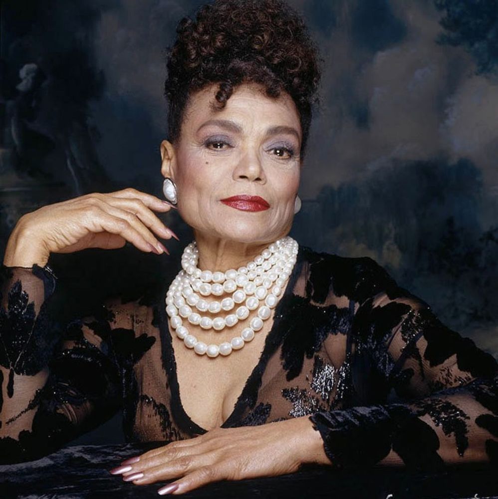 An elegant, sophisticated, and stunning portrait of Eartha Kitt in her later years. She is wearing a black lace gown, white pearl necklace, and red lipstick.