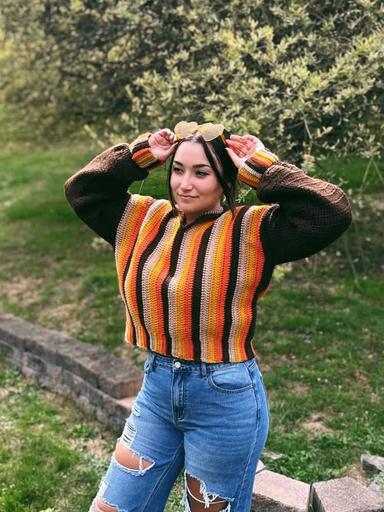 me outside wearing my striped crochet sweater with yellow heart shaped glasses on my head. I’m wearing jeans and my hands are up adjusting my sunglasses to show the cropped length of the sweater