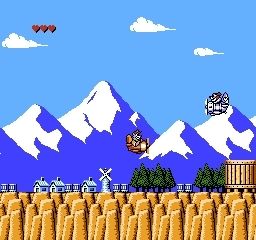 Screenshot of the Talespin game for Nintendo Entertainment System. It shows a tiny plane piloted by a talking bear from the cartoon, flying over small houses and a windmill.