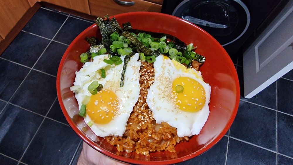 Kimchi fried rice (bokkeumbap) with toasted seaweed strips, spring onion, toasted sesame seeds and two sunny side up eggs, in a deep red bowl