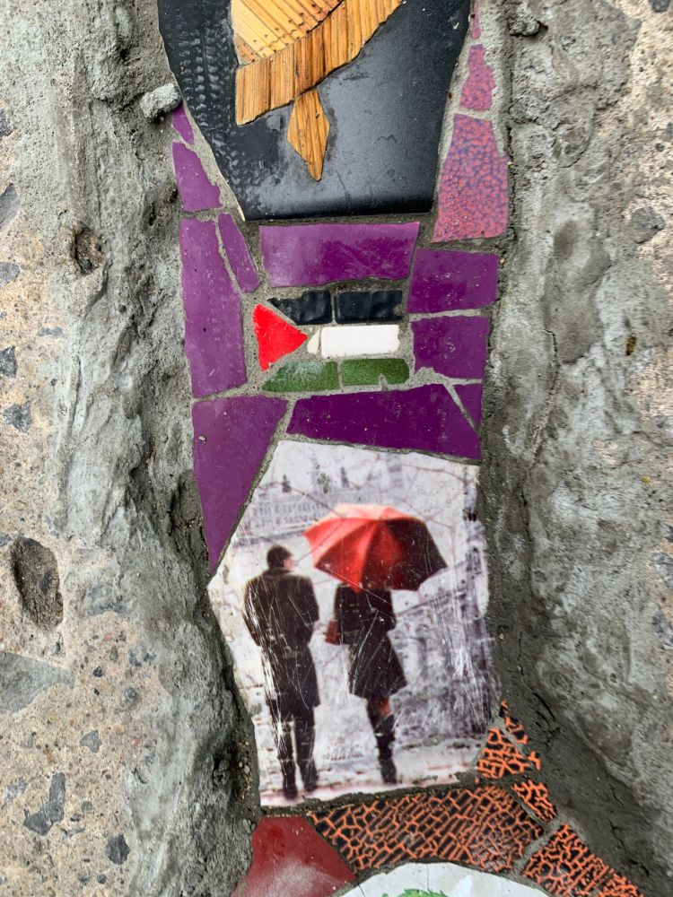 Close-up of tile work in crack featuring a tile of back-facing view of man and woman walking, woman carries a red umbrella, above this are green, white, black, and red tiles arranged to form the Palestinian flag.