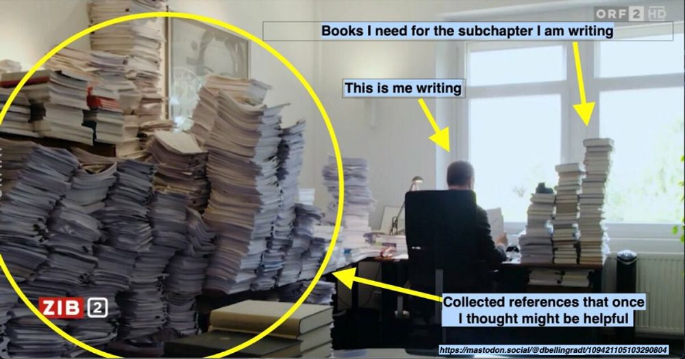 Photo of a working desk with loads of papers, and someone working at a laptop.