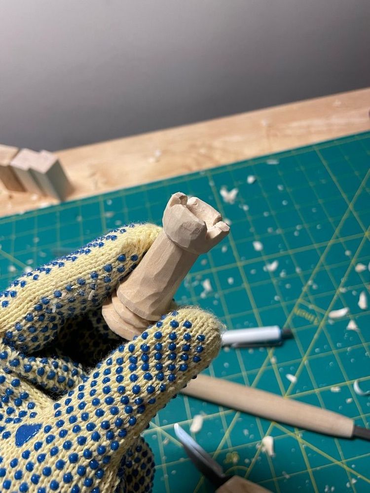 A kevlar gloved hand holds a small bit of wood that has been carved into the shape of a rook chess piece. In the background is a cutting mat with some tools on it and a bunch of wood shavings everywhere 