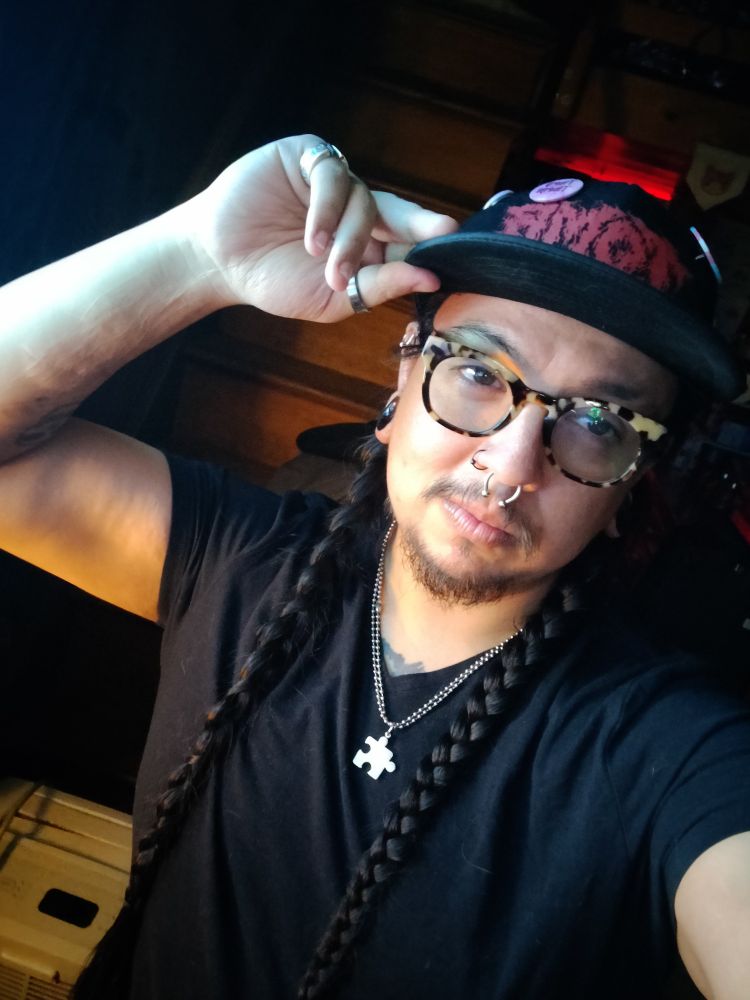 Johnny with a snapback on with some pin buttons on it, plastic rimmed glasses, hair braided into two braids. They're wearing a puzzle piece necklace, a black T-shirt, and has stretched ears and two nose rings. They're holding the brim of their hat with their right hand which has a ring on the thumb and pinkie finger. 