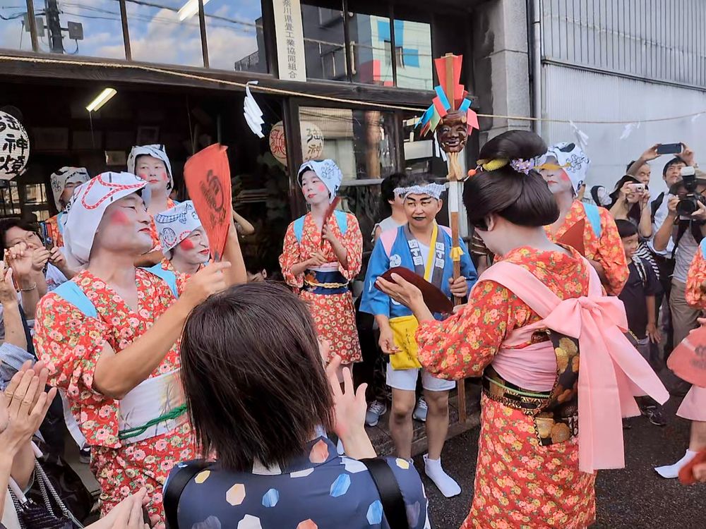 A group of men in red women's kimono get ready to distribute good health charms in front of an old shop.