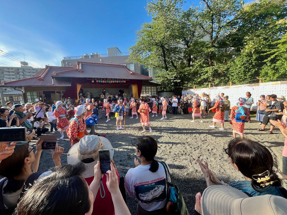 A group of men in red women's kimono dance in a circle at a Japanese shrine.