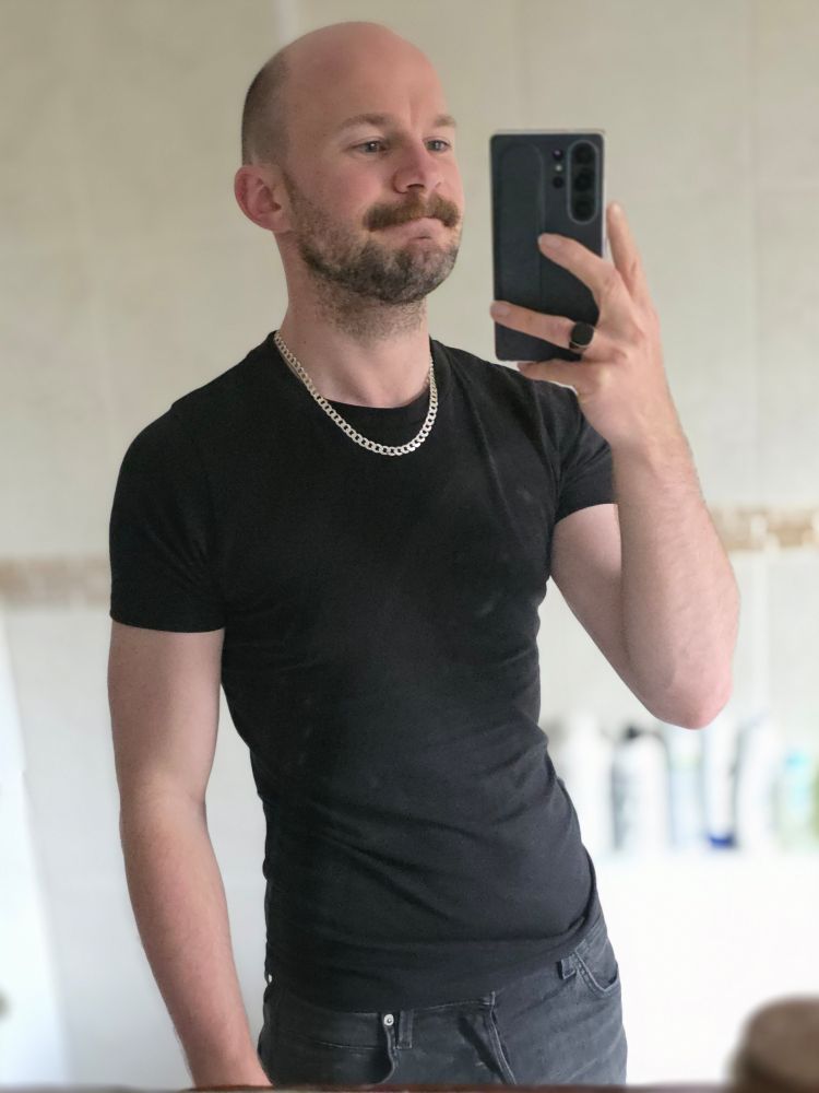 A man in a black t-shirt and shorts, wearing a silver chain necklace and black onyx ring is taking a picture of himself in the mirror