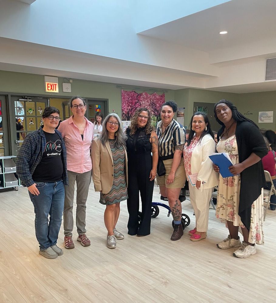 A group of 7 diverse people inside of a park district meeting room. From the left: Asher McMaher, a transmasc nonbinary person with light skin, dark curly hair, wearing a Trans Up Front Shows Up shirt with flannel over, blue jeans, and grey shoes. Kelly Cassidy, a light skinned woman with light skin and her hair in a ponytail, wearing a pink buttonup, grey pants, and brown shoes. Next is an asian woman with grey hair, a tan suit coat over a fun stripy dress and sneakers. Next is a curley haired woman wearing all black with light skin. Next is Silas, a transmasc person with light skin and curly dark hair wearing a black and white striped button down, tan shorts, and using a mobility scooter. Next is a darker skinned woman with dark brown hair, a white suit, with a pink shirt and heels. Last is a black woman with braids or locs holding a blue folder, wearing a floral light colored dress, black sweater, and sneakers. 
