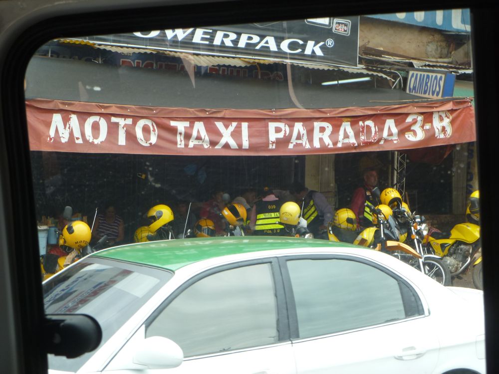 Die untere Bildhälfte wird von einem parkenden PKW verdeckt. Dahinter sieht man mehrere Motorräder, an deren Lenkern hellgelbe Motorrad hängen. Darüber ein Banner mit der Aufschrift Moto Taxi Parada.