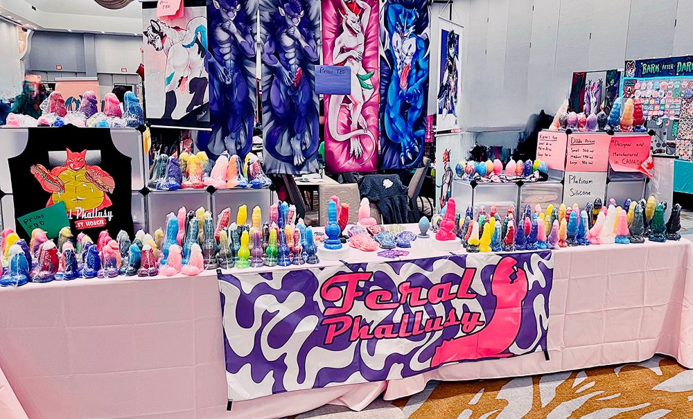 Photo of 18+ dealers den table for Feral Phallusy. Many dildos cover the table. A line up of our double sided dakis and wall scrolls stands behind it.