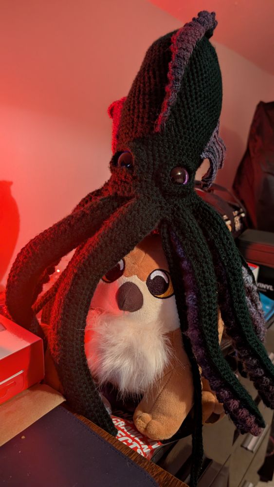 A green and purple crochet Kraken sits on top of a plush out there with red lighting from the left 