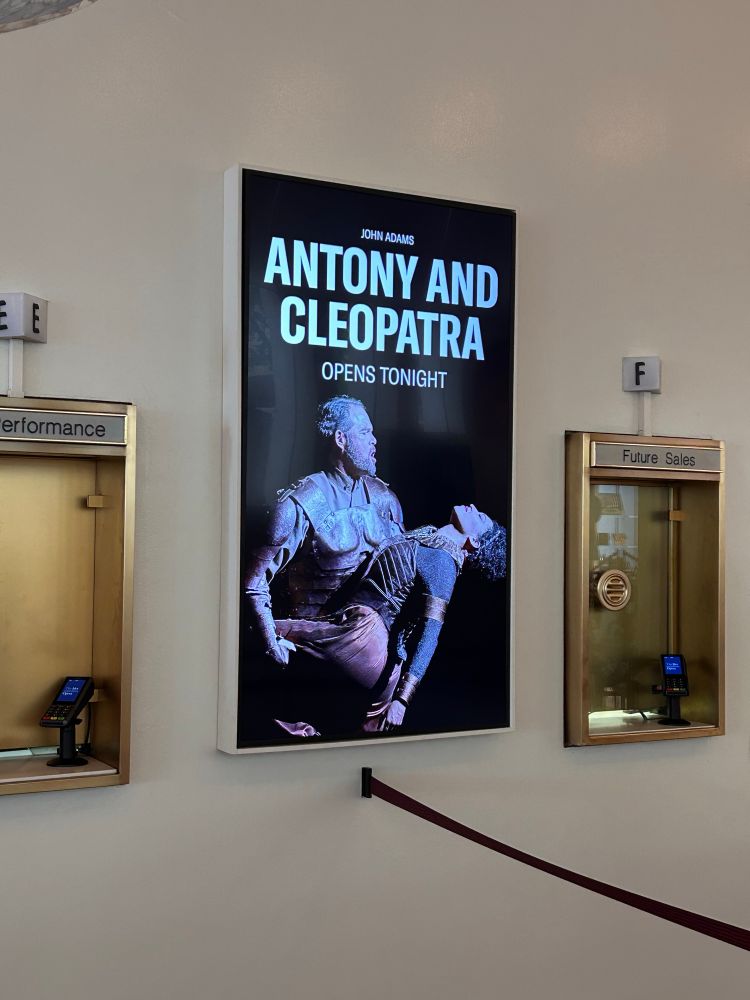 a digital poster at the Metropolitan Opera House box office that shows Gerald Finley as Mark Antony dipping back Julia Bullock as Cleopatra. it announces ANTONY AND CLEOPATRA OPENS TONIGHT
