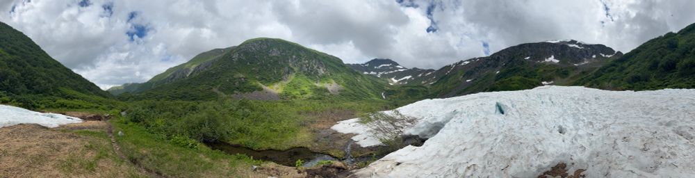 A pano shot of a valley with a pile of snow and green hills. 