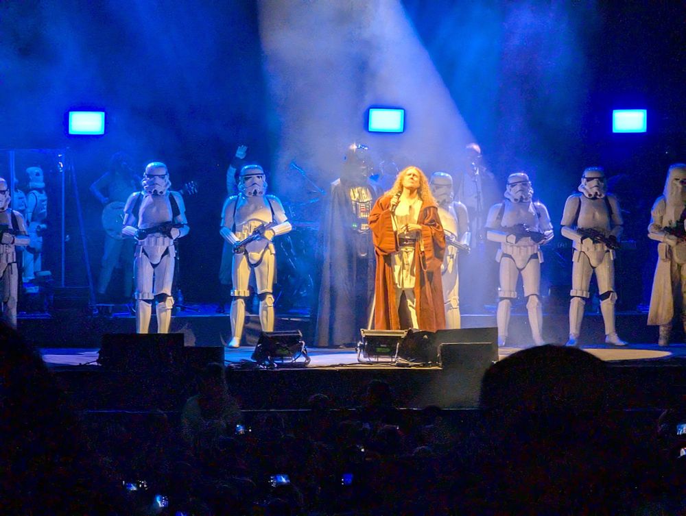Weird Al, dressed like a Jedi, spotlighted on stage in front of a row of Storm Troopers.