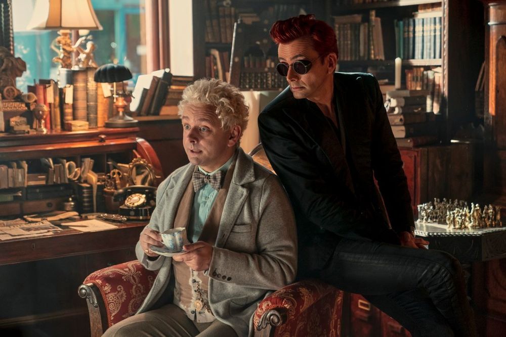 In Aziraphale's (Michael Sheen) bookshop. He (in worn suit) sits in a comfy chair, holding a cup of tea, whilst Crowley (David Tennant) sits on the arm, his back (clad all in black) against Aziraphale's shoulder. An old, delightful & cosy bookshop around them.