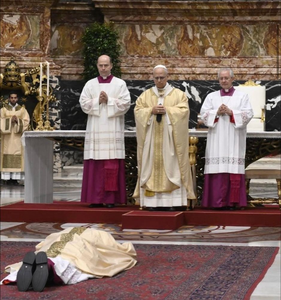 Na Basílica de São Pedro, o Papa Leão preside a ordenação episcopal de Dom Wachowski. Em primeiro plano, o ordenando está prostrado diante do altar em gesto de entrega total, vestindo paramentos dourados. Atrás dele, o Papa e dois prelados em túnica branca e roxa permanecem em oração solene diante do altar, cercados por outros clérigos.