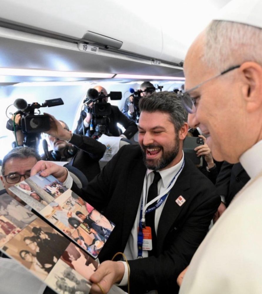 Jornalista branco de barba e cabelos castanhos mostra para Santidade um álbum cheio de fotos dele (Santidade) que olha com uma cara confusa entre felicidade e espanto