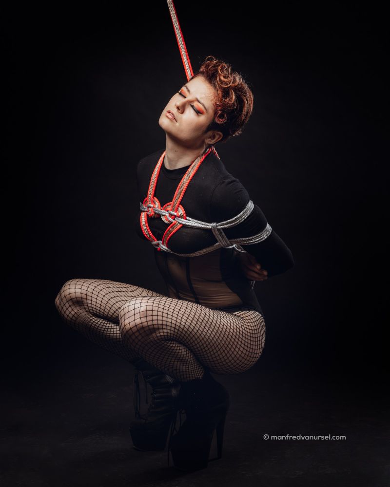 the girl is still squatted, upper body upright giving a view of the silver, red and golden heart harness. A rope still runs upwards out of frame from the back of the harness.