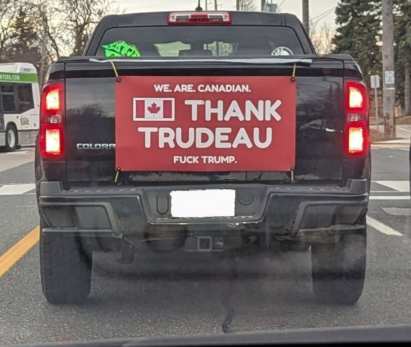 Back of a black pickup truck with a large red decal attached to the tailgate. In addition to a Canadian flag, the decal reads, "We are Canadian. Thank Trudeau, fuck Trump." All parties are stopped at a newly-turned red light and not behaving dangerously.