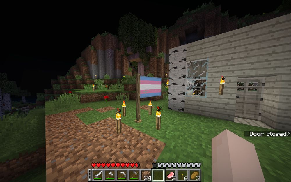 a trans pride flag in the BetterThanAdventure Minecraft mod, surrounded by torches. My house, built out of birch wood and lacking a proper roof is visible on the right.