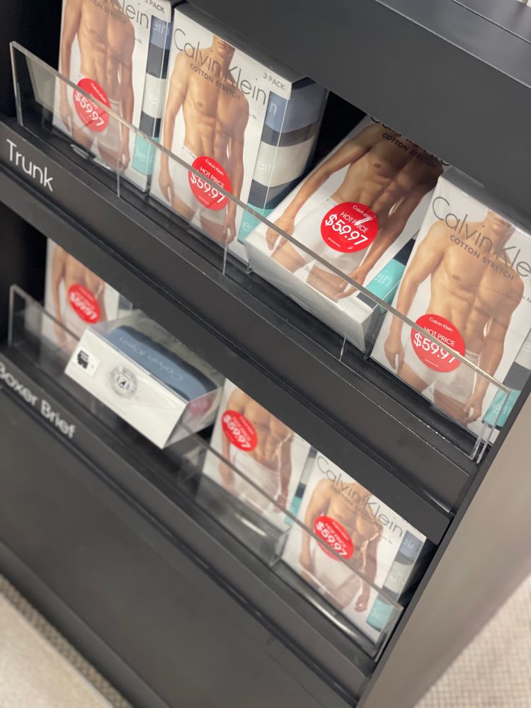 Photo of a display case of Calvin Klein Underwear boxes. Large red price stickers cover the model’s bulge on almost every box visible.