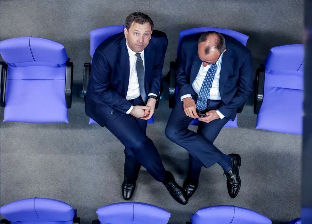 Lars Klingbeil und Friedrich Merz im Bundestag von oben fotografiert, sie sitzen nebeneinander, jeweils das rechte über das linke Bein geschlagen und identisch gekleidet