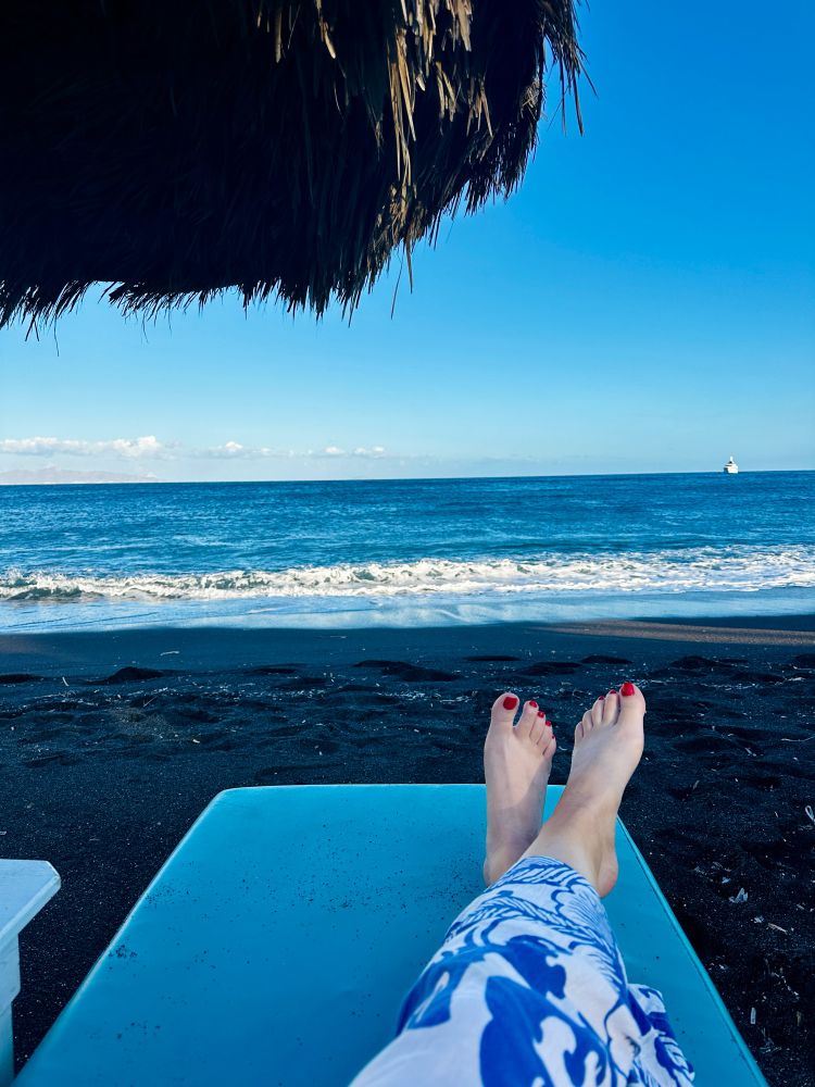 Shot of woman resting on a lounge chair on the beach, looking at the ocean and blue sky under a bamboo umbrella. Her legs are crossed and you can only see her feet. 