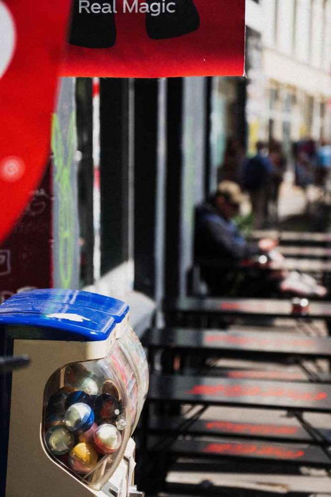 Photo showing a capsule machine and soda advertisements in the foreground with a man sitting in the background, out of focus.
