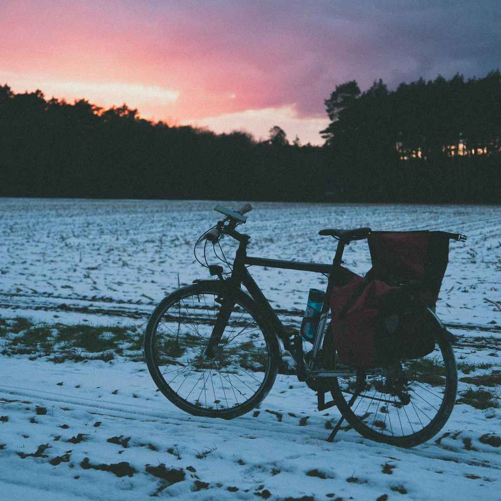Photo of a bike in the snow. In the background is a sunset behind trees.