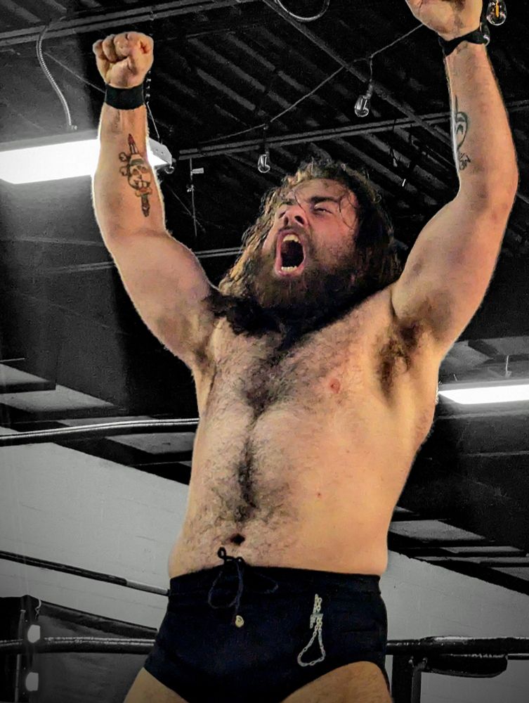 It's Mad Dog Connelly! He's a white man with long brown hair and brown beard wearing a pair of black wrestling trunks with an image of a chain shaped like a noose on the side. He has his fists raised in the air and is yelling with rage.