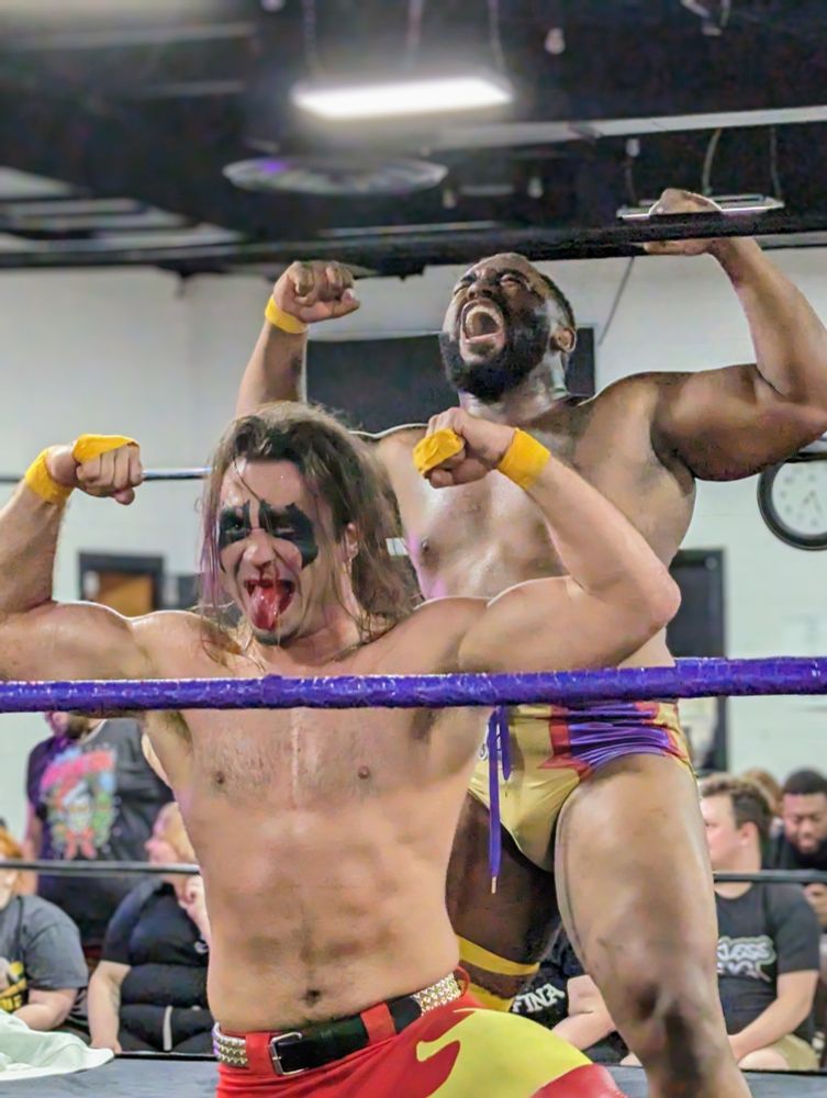 It's Camaro Jackson and Warhorse! Camaro is a black man and Warhorse is white. Both are wearing wrestling tights and are flexing to the crowd after a victory. Warhorse's tights are red and yellow pants while Camaro is wearing briefs that are purple and gold. Warhorse also has black face paint around his eyes.