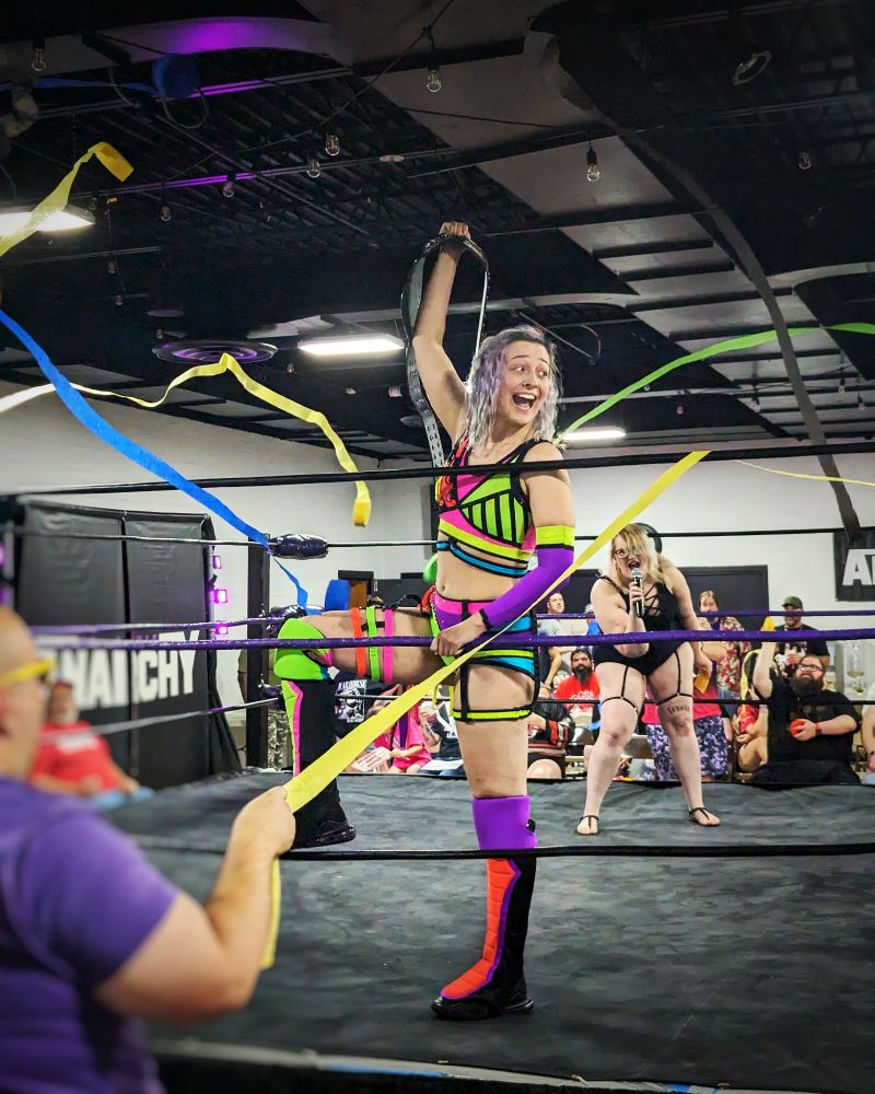 It's Billie Starkz! She's a Caucasian woman wearing pink and green wrestling tights, holding up her ROH Women's TV Title, and giving an expression of joy as multi-colored streamers fly around her. In the near background is High Voltage Kat, a blonde Caucasian woman, wearing an all-black outfit and screaming into the microphone as she introduces Billie to the crowd.