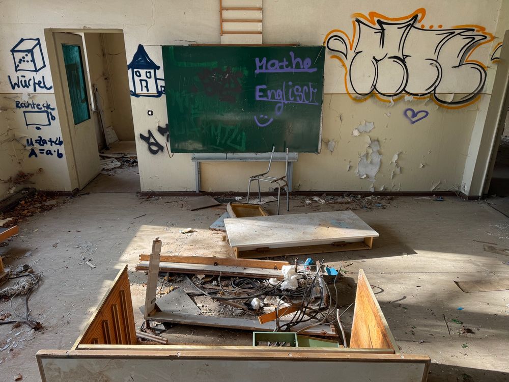 Ein verlassenes Klassenzimmer mit einer Tafel mit der Aufschrift "Mathe" und "Englisch", die verstreute Trümmer, zerbrochene Möbel und Graffiti an den Wänden zeigt. Das Sonnenlicht strömt durch und beleuchtet die Unordnung. Links ein gesprayter Würfel, darunter mathematische Formeln.