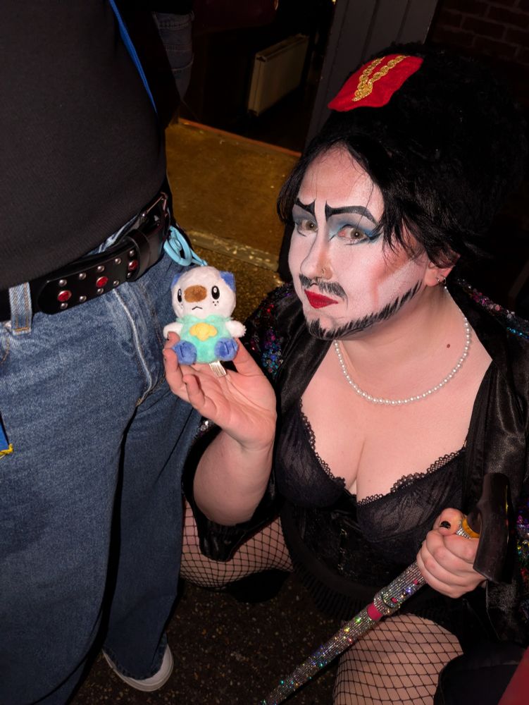 A picture of Viktor Complex (white drag king) with Dr Frank N Furter inspired white makeup posing with a small Oshawott plush hanging from a man's belt loop. 