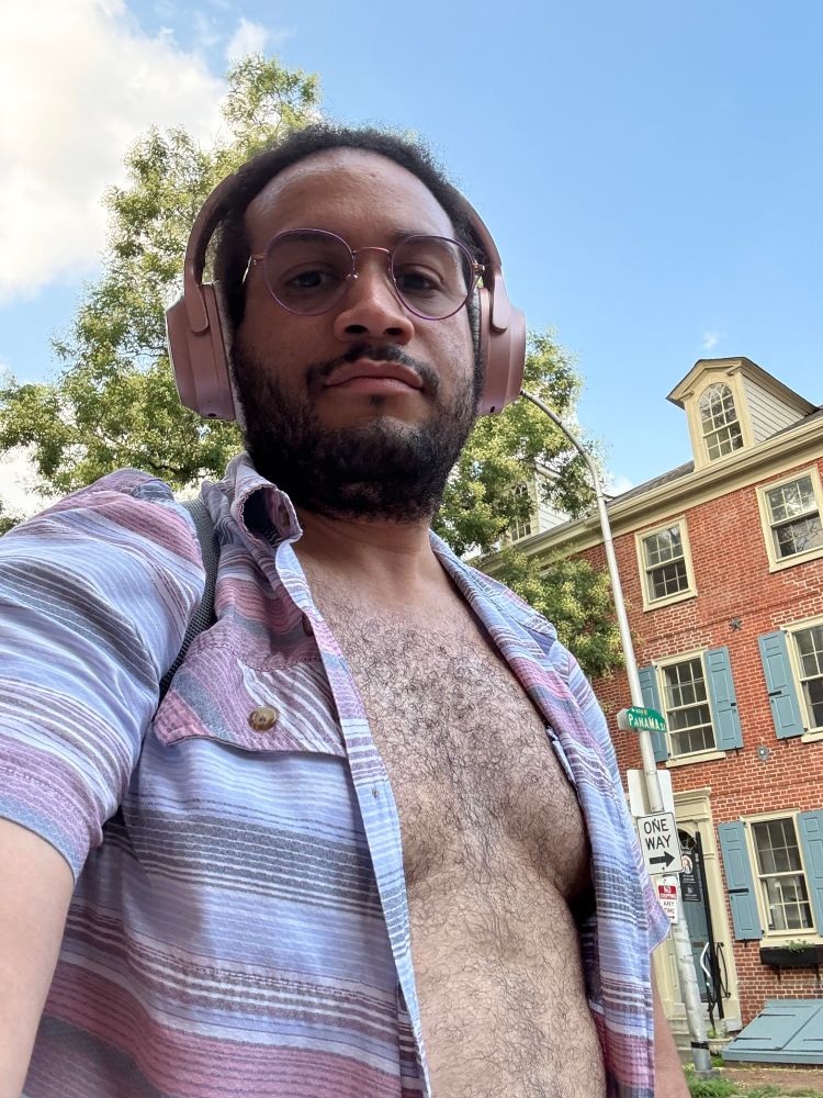 Selfie of Vega, enby with dark brown hair pulled back. They are wearing purple glasses and a pink set of over-the-ear headphones. They are wearing and opened button up shirt with various thin stripes of blue, black, red, white, and grey. Their chest is open with the shirt covering their nipples, copius chest hair. Vega is outdoors, a blue sky with a white cloud behind a large tree. There's a colonial-style red brick house with light blue windows. They are looking at the camera with a very neutral expression. 
