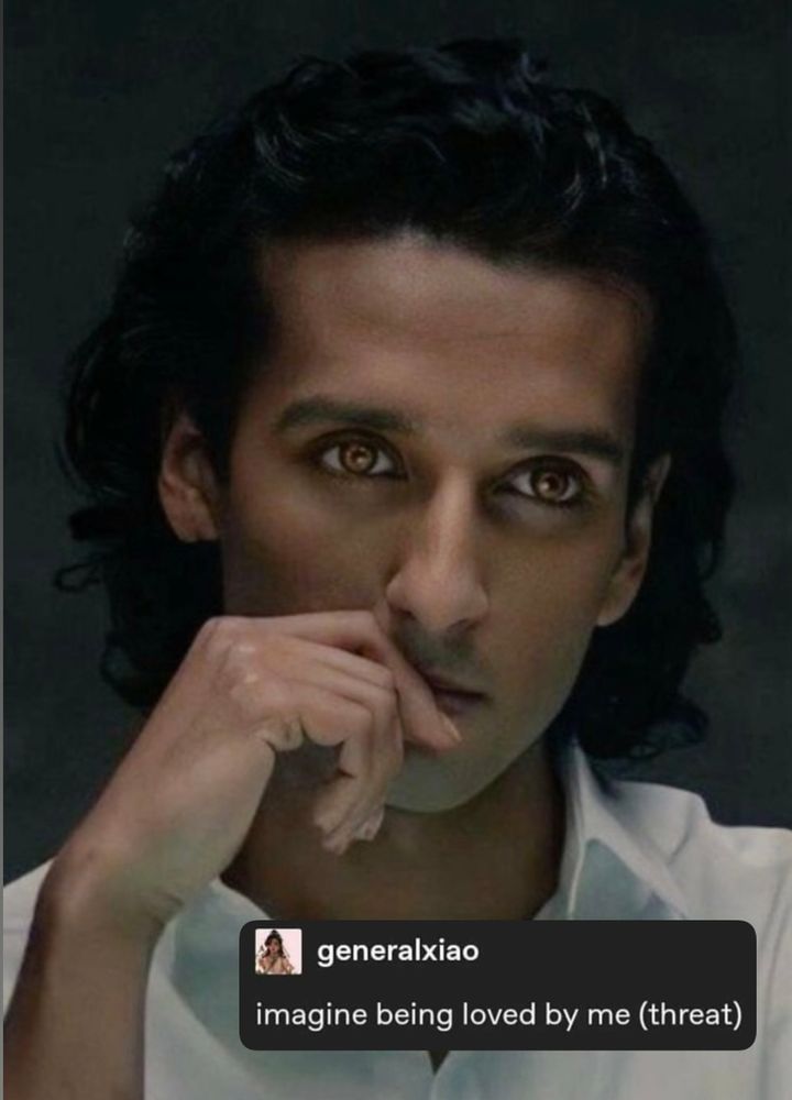 A closeup of Armand from interview with the vampire (2022), played by Assad Zaman. He’s wearing a white shirt with the top buttons undone. One hand rests on his chin, half covering his lips. He’s looking intensely at someone out of frame. I have added a screenshot of a tweet that reads: “imagine being loved by me(threat).” 