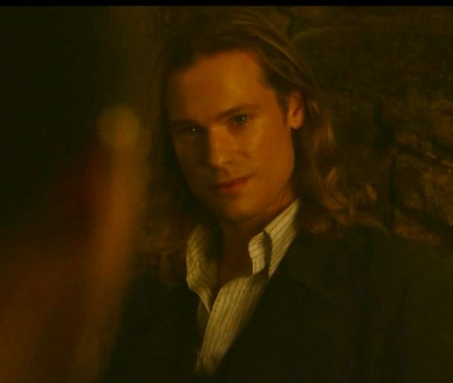 Lestat in the dungeon after the trial. He’s standing against a dark background, wearing a light striped and collared shirt and a dark coat. His hair is down, and he’s looking toward someone with a small, exhausted smirk. 