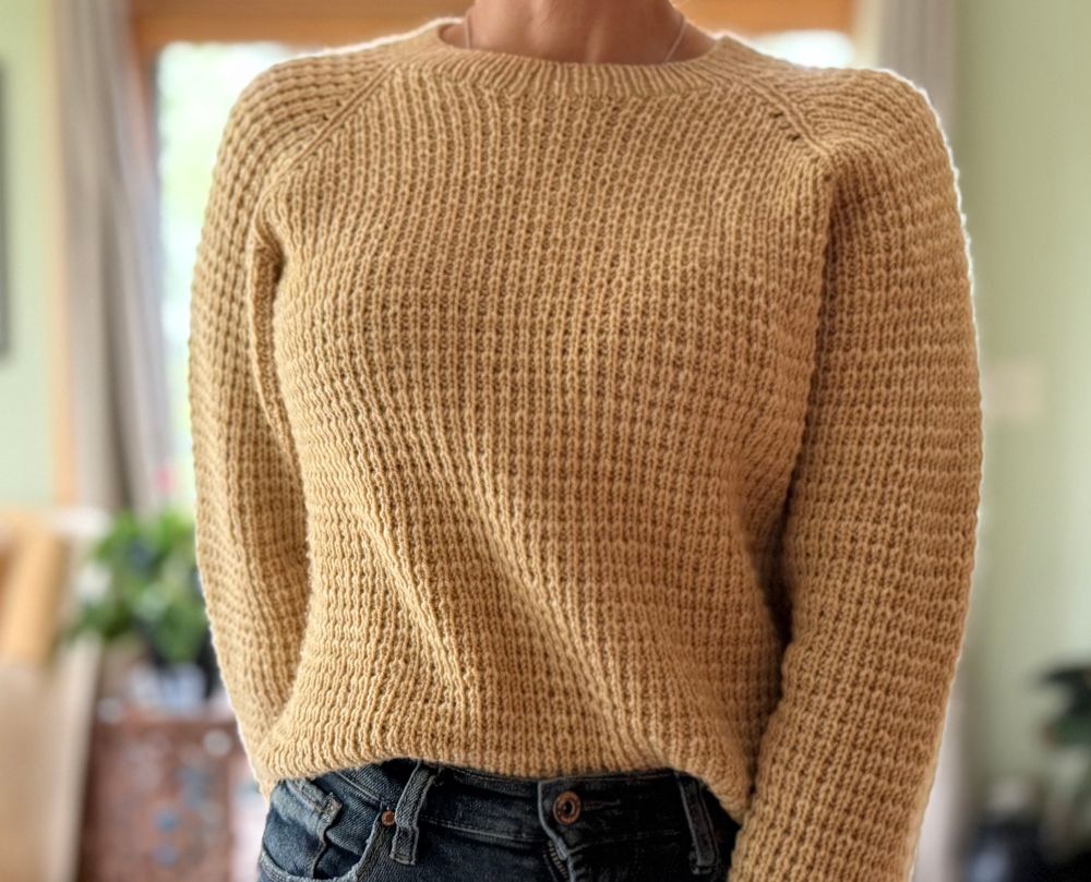 The knitter modeling a hand knit sweater with a Fisherman’s rib stitch pattern in wheat colored yarn. The pattern is Mabel’s Sweater by Joji Locatelli and the yarn is Purl Soho Wigeon in an unnamed sample color.