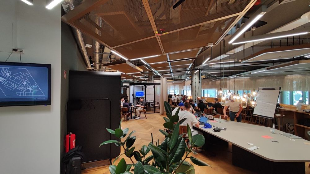 The interior of EDGE coworking spaces in Berlin, showing different teams of people sitting around tables and working together during the 2025 hackathon by 50hertz. Several screens and flipcharts are visible. The coworking space has a cosy atmosphere with wooden floors, plants, and organically shaped furniture.